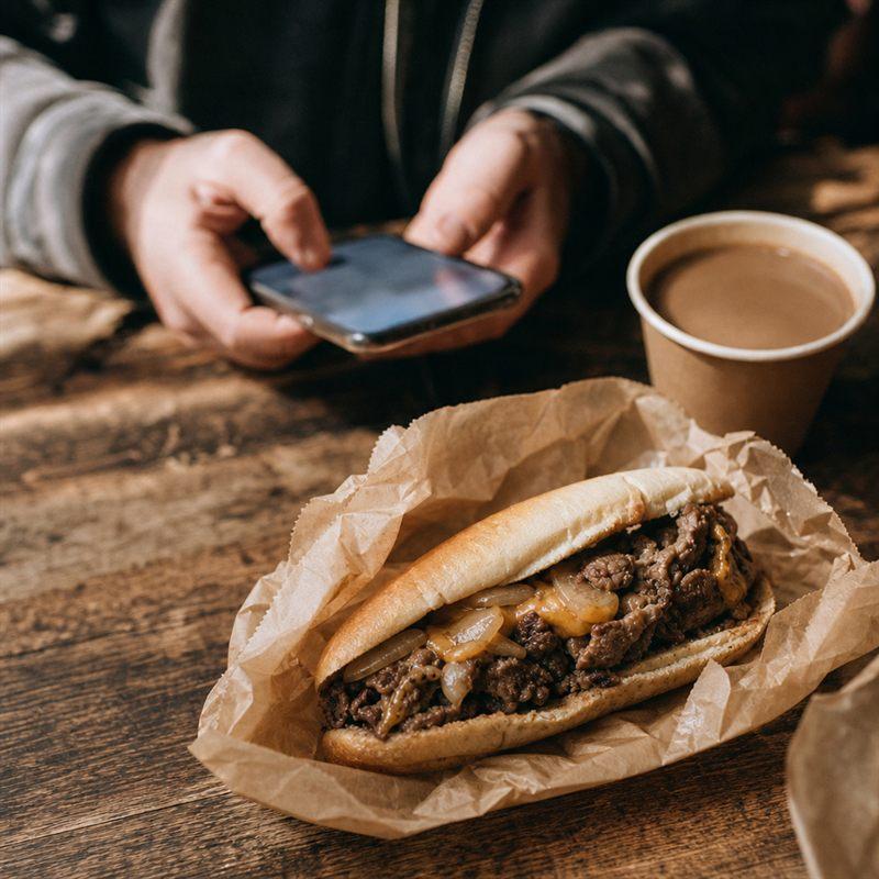 Hand am Smartphone neben einem frisch gegrillten Philly Cheesesteak
